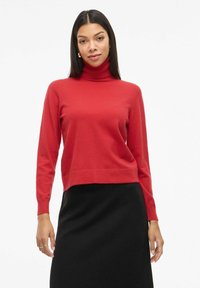 Crvena turtleneck pulover s dugim rukavima i skraćenim dizajnom. Napravljen od mekanog materijala, s rebrastim izrezom i manžetama. Ispod se nalazi crna suknja.