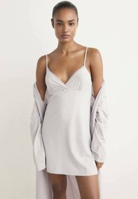 Femme aux cheveux tirés en arrière portant une robe nuisette légère lavande à fines bretelles, avec un manteau assorti drapé sur ses bras, sur un fond uni.
