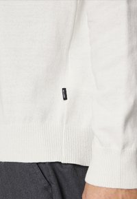 Maglione bianco a maglia con polsini e orlo a costine, dotato di un piccolo etichetta nera sul lato. Il tessuto ha una consistenza fine e un aspetto leggero.