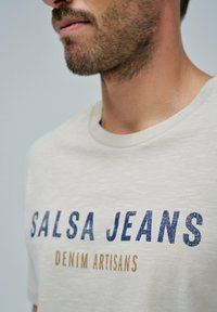 Camiseta beige con estampado texturizado azul que dice "SALSA JEANS" y "DENIM ARTISANS" en una fuente más pequeña debajo. Cuello redondo, tela suave.