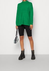 Grüner Strickpullover mit hohem Kragen, kombiniert mit schwarzen Radlerhosen und klobigen schwarzen Schnürboots. Die Tasche hat ein Schwarz-Grünes Muster.