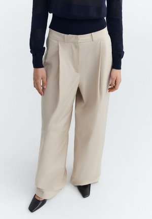 Woman wearing beige wide-leg trousers, black crewneck sweater, and black pointed-toe shoes standing against a white background.