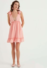 Rosa ärmelloses Kleid mit tiefem V-Ausschnitt, schwingendem Saum und Schleifenakzenten an den Schultern. Der Stoff hat eine subtile Textur und ein Pünktchenmuster.
