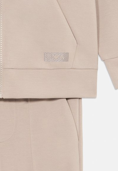 Beige Kapuzenpullover mit Reißverschluss, der ein strukturiertes Logo-Patch, eine Kapuze und große Fronttaschen zeigt. Weicher Stoff mit einer glatten Textur und minimalistisches Design.