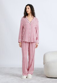 Γυναίκα που στέκεται φορώντας ροζ και λευκή πυτζάμα με καρώ σχέδιο και λευκές παντόφλες, δίπλα σε μια λευκή υφασμάτινη πολυθρόνα πάνω σε γκρι χαλί.