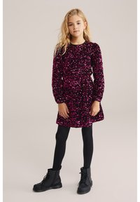 Rochie roz strălucitoare din paiete, cu mâneci lungi și fustă evazată; asortată cu colanți negri și cizme de luptă negre. Textură de țesătură netedă.