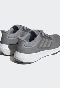 Scarpe sportive grigie con una parte superiore in maglia strutturata, dettaglio a tre righe e una suola intermedia bianca. Forma arrotondata con colletto imbottito e lacci.