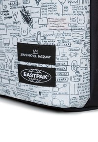 Reppu, jossa on vaaleanharmaa kangas mustilla graafisilla sketseillä, kaksi mustaa etikettiä, joissa on "JEAN-MICHEL BASQUIAT" ja "EASTPAK" -logot.