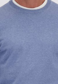 Lichtblauwe ronde halstrui van zacht gebreid materiaal. Heeft een eenvoudig ontwerp met een subtiele logo-detail nabij de halslijn.