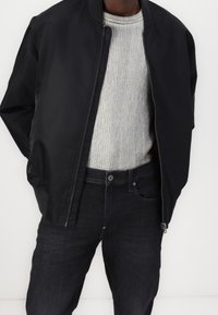 Chaqueta bomber negra con un acabado suave, cierre de cremallera y bolsillos laterales, combinada con un suéter de canalé gris claro y jeans de mezclilla oscura.