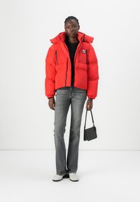 Red cropped puffer jacket with elastic cuffs and two front pockets, paired with gray flared jeans and white shoes, holding a small black bag.