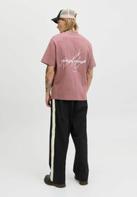 Rosa bomulds T-shirt med et hvidt grafiktryk på ryggen. Kombinationen med sorte bukser, der har en hvid sidestribe, og mørkt fodtøj.