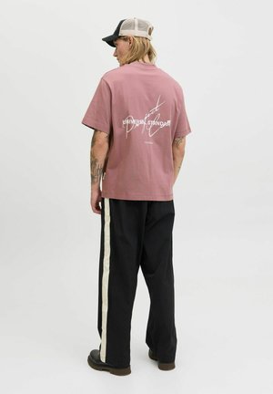 Rosa bomulds T-shirt med et hvidt grafiktryk på ryggen. Kombinationen med sorte bukser, der har en hvid sidestribe, og mørkt fodtøj.
