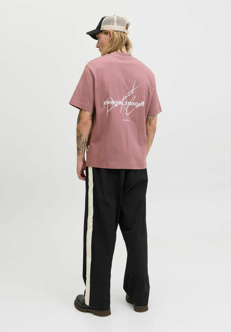 Rosa bomulds T-shirt med et hvidt grafiktryk på ryggen. Kombinationen med sorte bukser, der har en hvid sidestribe, og mørkt fodtøj.