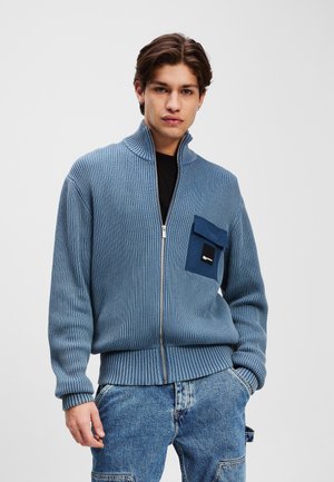Blå ribbet jakke med lynlås og høj krave, med en mørkeblå lomme med et label-detail, parret med lyseblå denimbukser.
