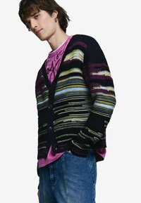 Pletený sveter so sivomodrými, fialovými a žltými vodorovnými pruhmi, čiernymi gombíkmi a rebrovanými manžetami. Nosený cez ružové tričko.