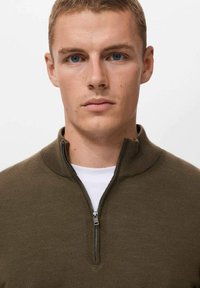 Suéter de cremallera color verde oliva con cuello alto y una cremallera plateada, usado sobre una camisa blanca. Textura suave, diseño ajustado.