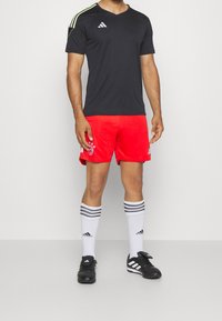 Maglietta atletica nera a scollo a V con dettagli verdi, pantaloni corti rossi con un logo, calze bianche con strisce nere e scarpe da calcio nere.
