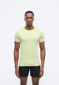 Homme debout face à l'avant, portant une chemise ajustée à manches courtes vert clair et un short noir, sur un fond blanc uni.