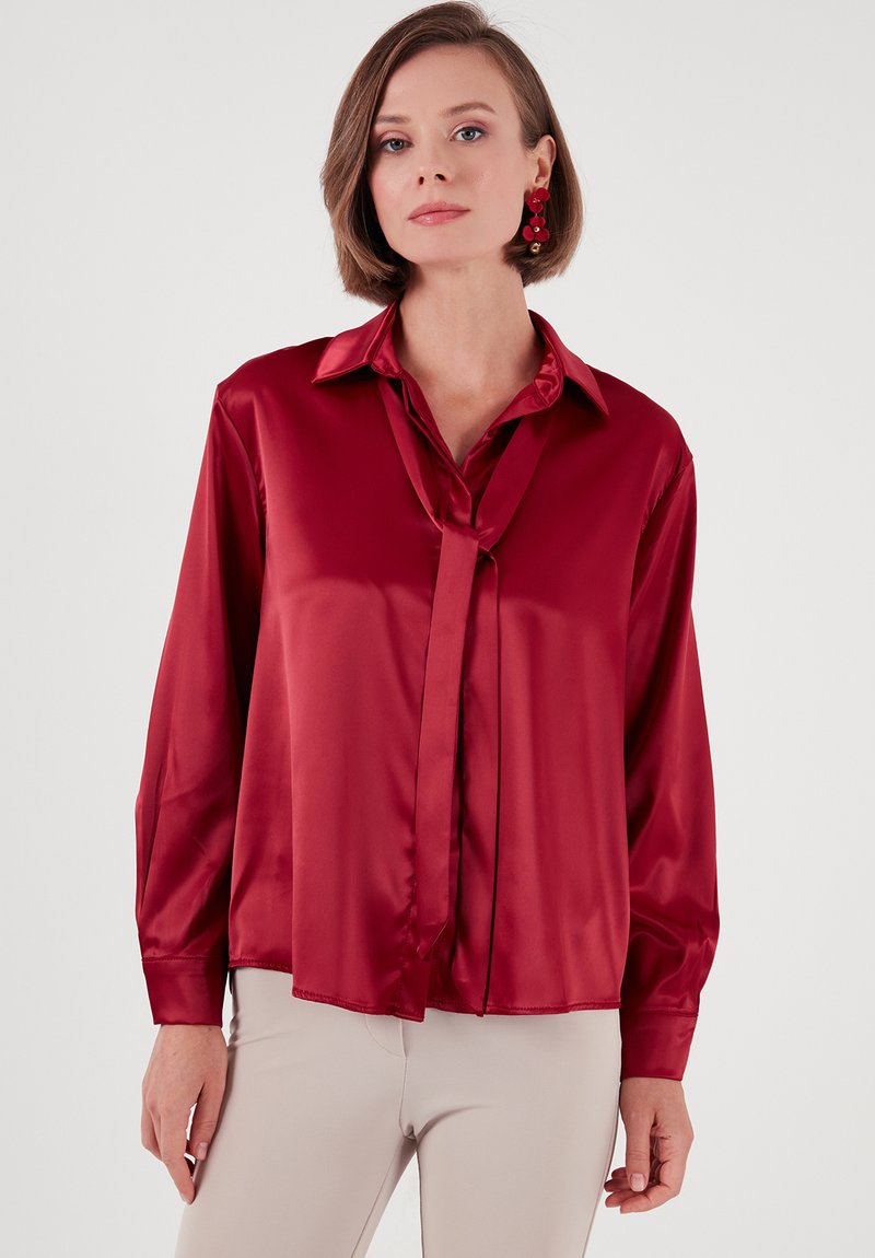 Red satin blouse with a classic collar, long sleeves, and a necktie detail. Smooth texture and relaxed fit. No visible patterns or hardware.