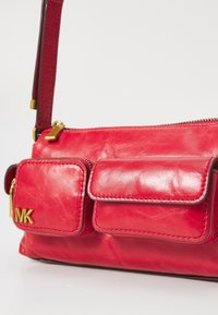 Rote Lederhandtasche mit Reißverschluss, ausgestattet mit zwei Vordertaschen, goldenen Beschlägen und einem abnehmbaren Tragegurt. Strukturierte Oberfläche mit Nähdetails.