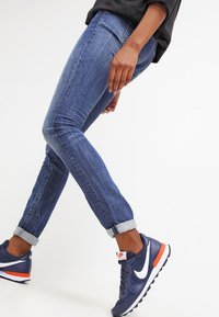 Osoba w podwiniętych niebieskich dżinsach i granatowych trampkach Nike z białym logo swooshem i pomarańczowymi akcentami spacerująca na białym tle.