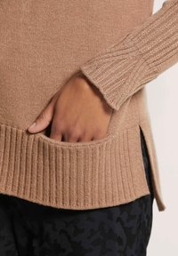 Beige gestrickter Pullover mit geripptem Saum und Bündchen, ausgestattet mit einem seitlichen Schlitz und einer Tasche auf Hüfthöhe, präsentiert mit einer Hand in der Tasche.