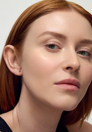 Złoty heksagonalny kolczyk na uchu modelki, posiadający gładką metaliczną powierzchnię. Modelka ma proste czerwone włosy i naturalny makijaż.