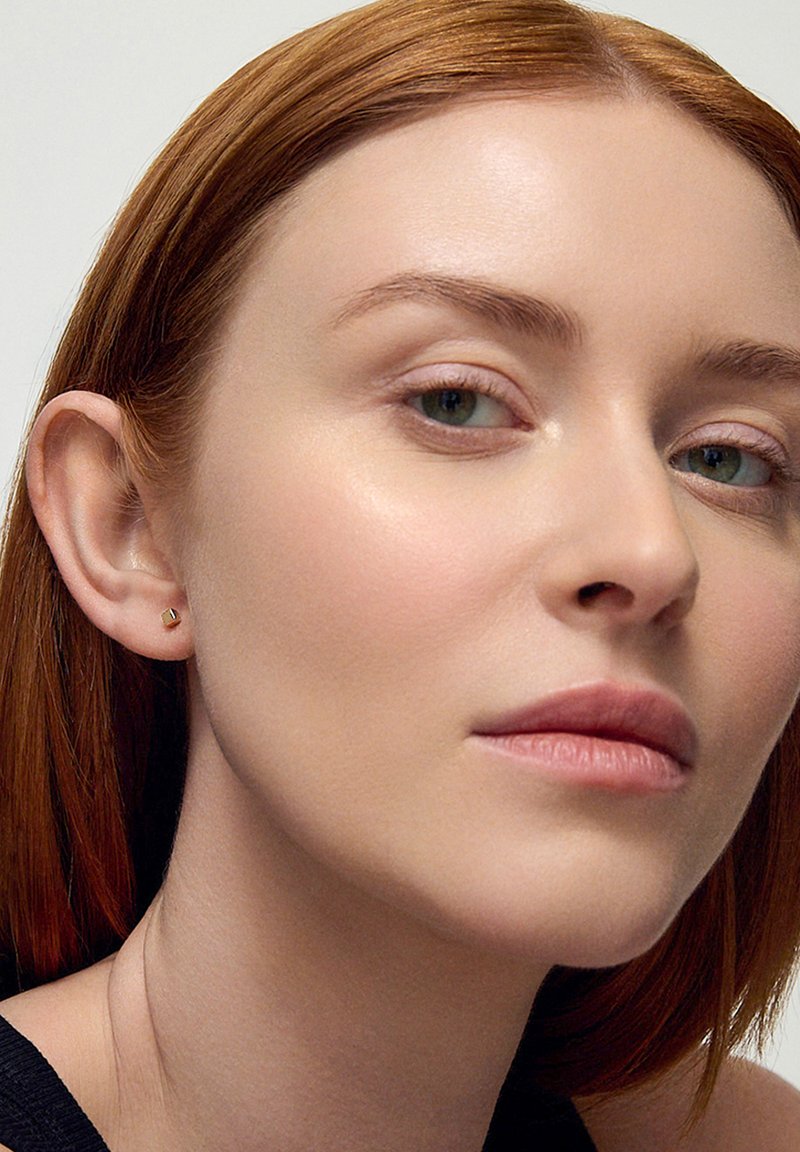 Złoty heksagonalny kolczyk na uchu modelki, posiadający gładką metaliczną powierzchnię. Modelka ma proste czerwone włosy i naturalny makijaż.