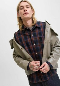 Grau leatherjacke, die über einem dunkelkarrierten Hemd mit blauen, braunen und weißen Quadraten getragen wird. Die oberen Knöpfe sind sichtbar; die Hände sind mit Tattoos verziert.