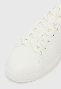 Sapatilha branca em couro sintético com superfície texturizada, atacadores brancos planos com fio dourado e bico arredondado. Design minimalista.