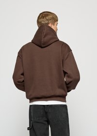 Brauner Hoodie mit großer Kapuze, lockerer Passform und gerippten Bündchen. Tragen über einem weißen Shirt, kombiniert mit dunklen Jeans. Weicher Stoff.