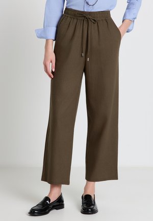 Donna con pantaloni a gamba larga a vita alta verde oliva con coulisse in vita e mocassini neri, in piedi davanti a uno sfondo semplice.