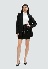 Schwarze Blazer über einem weißen Rollkragenpullover, kombiniert mit einem schwarzen Rock mit silbernen floralen Verzierungen. Schwarze High Heels mit dekorativen Akzenten.