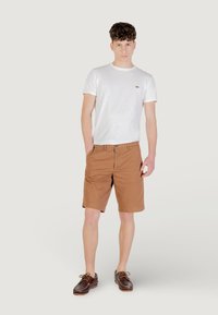 Shorts en coton marron et t-shirt blanc à col rond avec un petit logo vert. Le modèle porte des chaussures bateau marron. Arrière-plan neutre.