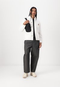 White canvas jacket, black shirt, and gray wide-leg pants. Person holds a black crossbody bag; wearing cream sneakers. Neutral background.