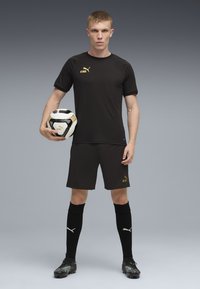 Sort atletisk trøje med gyldne accenter, matchende shorts, sorte sokker og fodboldstøvler. Spiller holder en sort-hvid fodbold.