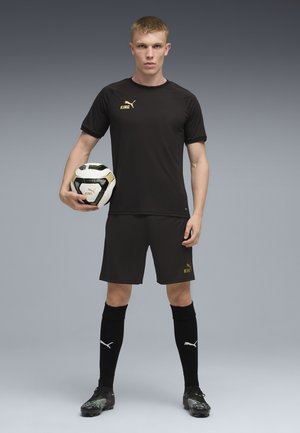 Sort atletisk trøje med gyldne accenter, matchende shorts, sorte sokker og fodboldstøvler. Spiller holder en sort-hvid fodbold.