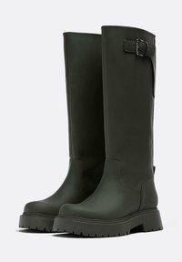 Knee-high, tmavě zelené gumové boty s hladkým povrchem, širším podpatkem a páskovým detailem na horní části. Disponují robustní podrážkou s drážkami.