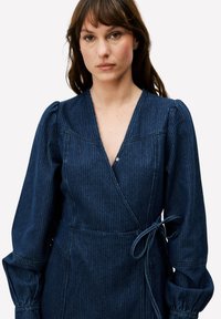 Robe portefeuille en denim foncé à rayures verticales, col en V profond, manches bouffantes et taille nouée. Détail de boutons près de l'encolure.