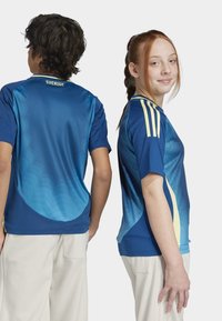 Sportovní dresy s modrým gradientem a žlutými akcenty, na rukávech tři pruhy. Krátký rukáv, lehký, prodyšný materiál s texturou síťoviny.