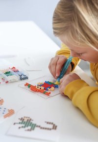 Perler kraal activiteit kit met kleurrijke kralen op een doorzichtige pegboard. Een kind gebruikt een blauwe tool voor het plaatsen. Patroonbladen liggen erbij.