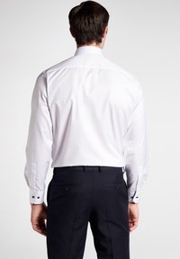 Camicia bianca a maniche lunghe con polsini a bottone, abbinata a pantaloni navy con doppi taschini posteriori e dettagli in bottone neri.