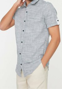 Camisa de botones de manga corta en gris claro con un patrón texturizado, botones azul marino, un bolsillo en el pecho y detalle en los puños.