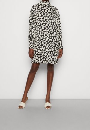 Robe florale noire avec des fleurs blanches, col haut, manches longues, design à mi-genoux, associée à des mules ouvertes beiges sur un fond gris.