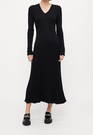 Femme portant une longue robe noire en maille côtelée avec un col en V et des manches longues, associée à des mocassins noirs ornés d'une chaîne dorée.