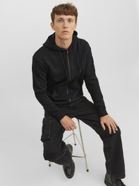 Juodas zip-up megztinis su kapuce, derinamas su juodomis cargo kelnėmis. Sklandus tekstūros paviršius su siūlėmis ir laisva mase. Derinamas su juodais batais.