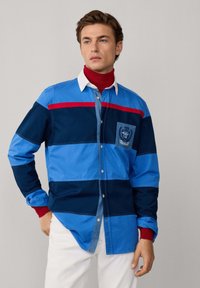 Blauw en marine gestreepte shirt met een witte kraag en rode accenten, met een borstzak met een embleem en knoopsluiting.