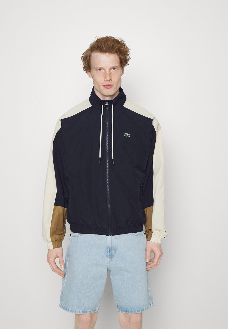 Marineblauwe en crèmekleurige windbreakerjas met een elastische tailleband, voorzien van een opstaande kraag, trekkoorden en contrasterende bruine accenten.