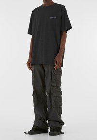 Camiseta negra de gran tamaño con un detalle gráfico, combinada con pantalones cargo negros que presentan múltiples bolsillos y un puño ajustado. Zapatillas negras completan el look.